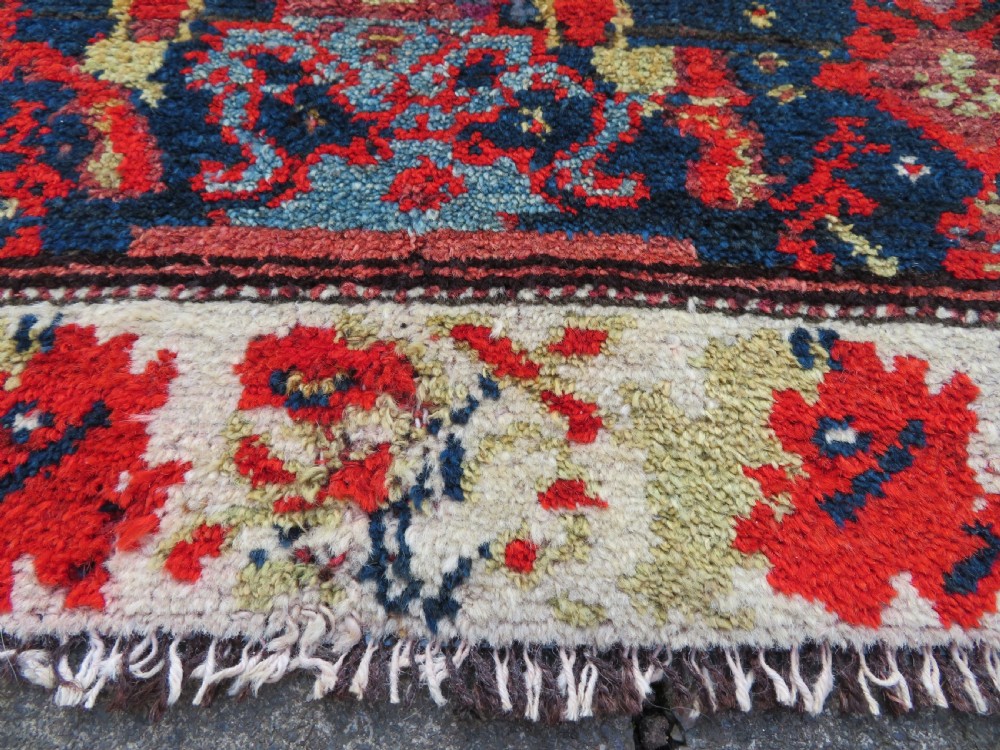 A rectangular hand-knotted antique Kurdish wool rug featuring red, navy blue, and cream tribal motifs with a decorated floral border. View XVIII
