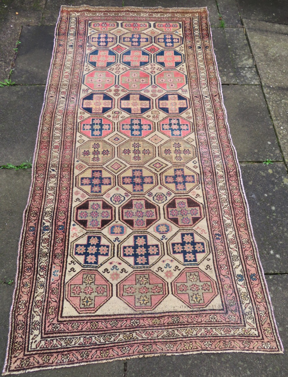An antique Kurdish hand-knotted wool rug featuring octagonal geometric medallions, cross motifs, stylized animal figures, and a multi-banded border in rose, beige, and navy.