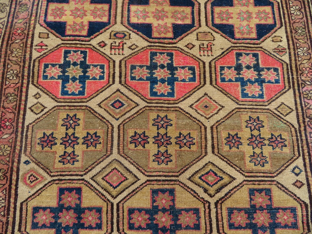 An antique Kurdish hand-knotted wool rug featuring octagonal geometric medallions, cross motifs, stylized animal figures, and a multi-banded border in rose, beige, and navy. View X