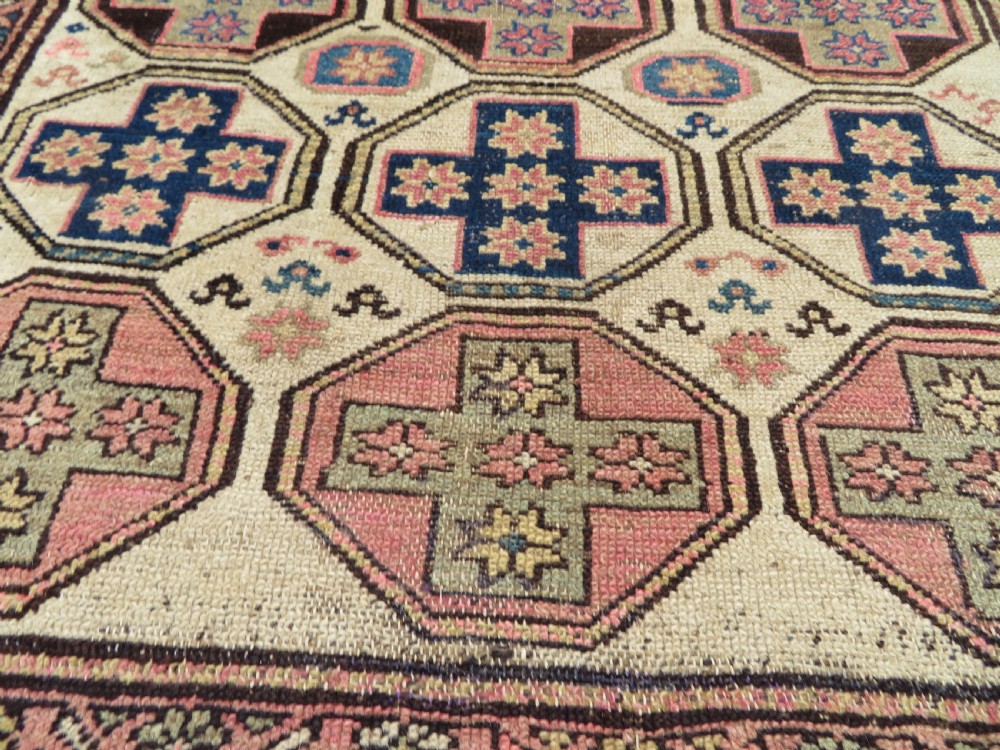 An antique Kurdish hand-knotted wool rug featuring octagonal geometric medallions, cross motifs, stylized animal figures, and a multi-banded border in rose, beige, and navy. View XX