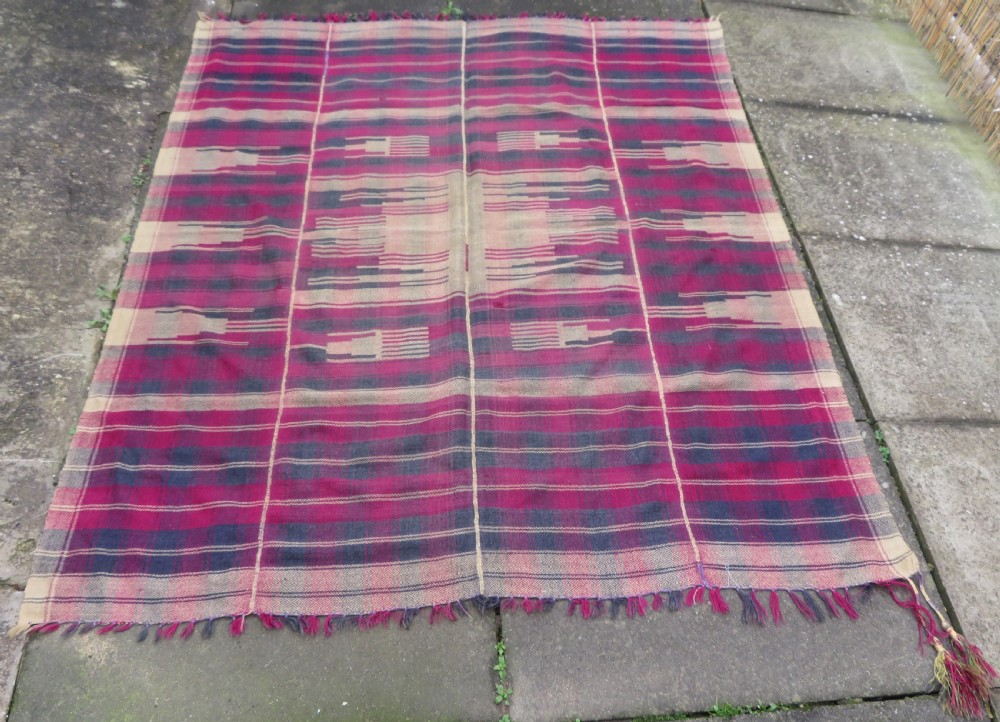 A rectangular flat-woven wool textile featuring crimson, navy, and cream geometric checked patterns with fringed ends.