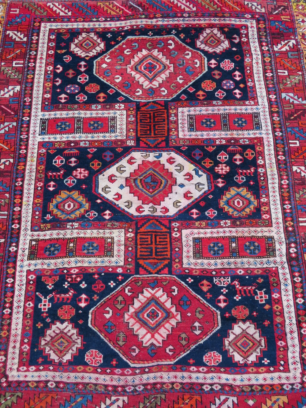 An antique late 19th-century Caucasian Kuba Konagkend wool rug featuring red geometric medallions set against a dark navy field with multi-coloured borders. View V