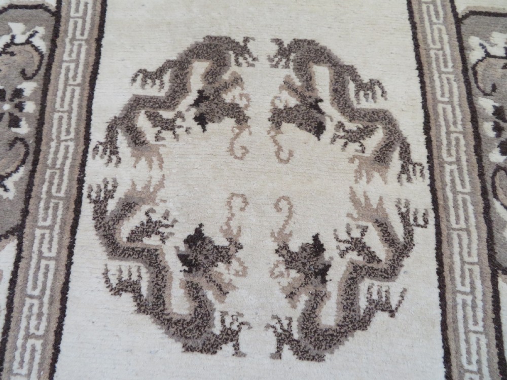 A rectangular hand-knotted cream wool rug featuring two central dragon medallions, a key meander border, and grey decorative motifs. View V