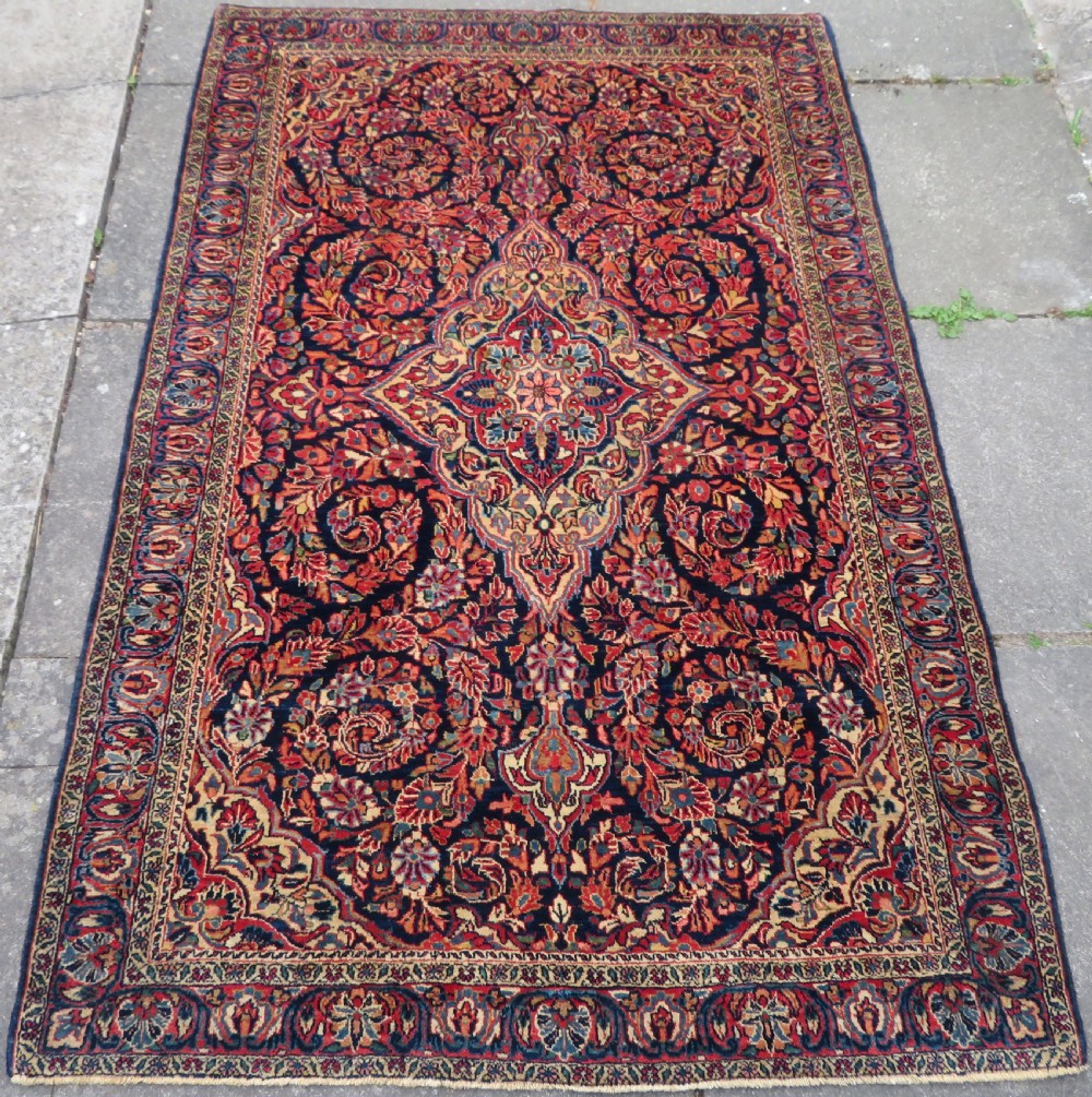 An antique West Persian Sarouk wool rug featuring a detailed central floral medallion framed by dense floral vine scrolls in shades of red, dark navy, cream, and blue.