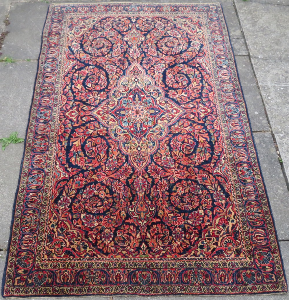 An antique West Persian Sarouk wool rug featuring a detailed central floral medallion framed by dense floral vine scrolls in shades of red, dark navy, cream, and blue. View II