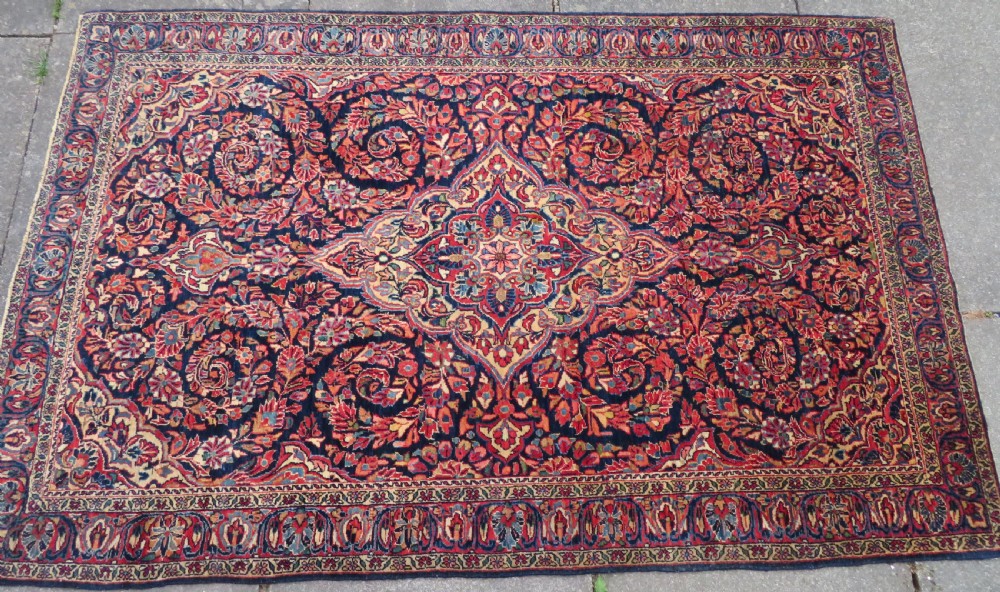 An antique West Persian Sarouk wool rug featuring a detailed central floral medallion framed by dense floral vine scrolls in shades of red, dark navy, cream, and blue. View III