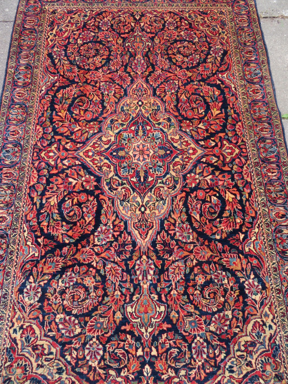 An antique West Persian Sarouk wool rug featuring a detailed central floral medallion framed by dense floral vine scrolls in shades of red, dark navy, cream, and blue. View IV
