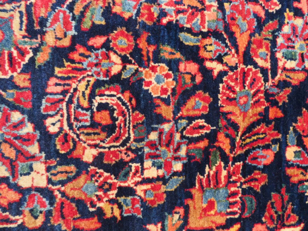 An antique West Persian Sarouk wool rug featuring a detailed central floral medallion framed by dense floral vine scrolls in shades of red, dark navy, cream, and blue. View X