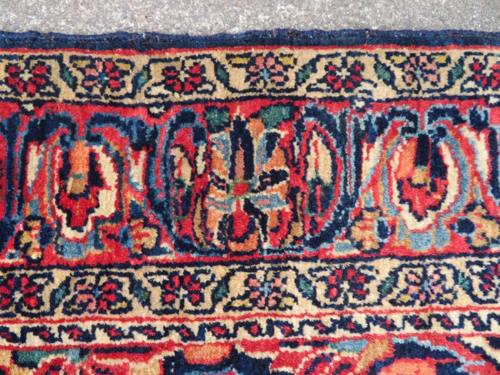 An antique West Persian Sarouk wool rug featuring a detailed central floral medallion framed by dense floral vine scrolls in shades of red, dark navy, cream, and blue. View XI