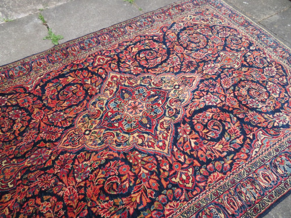 An antique West Persian Sarouk wool rug featuring a detailed central floral medallion framed by dense floral vine scrolls in shades of red, dark navy, cream, and blue. View XVI