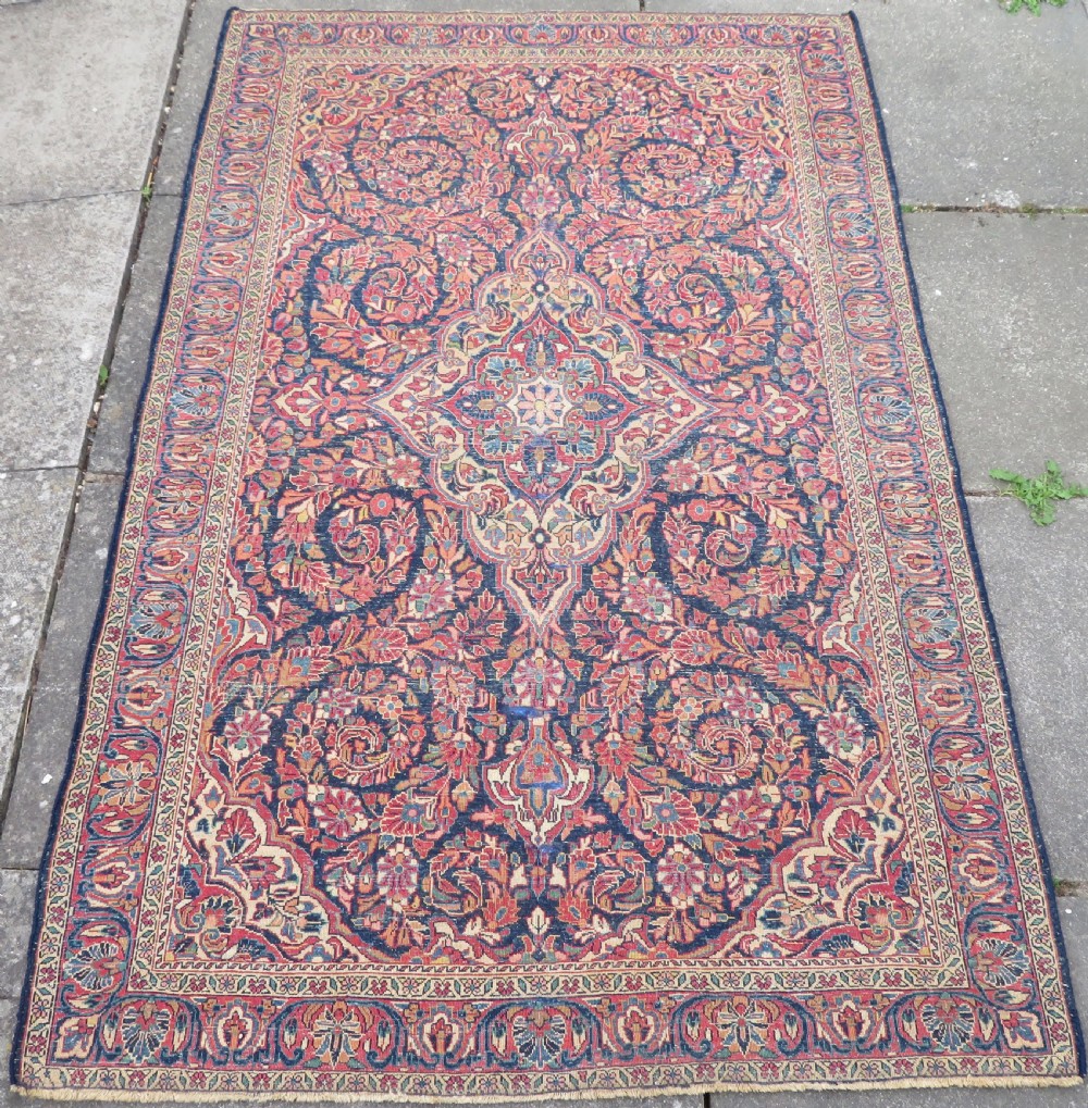 An antique West Persian Sarouk wool rug featuring a detailed central floral medallion framed by dense floral vine scrolls in shades of red, dark navy, cream, and blue. View XXI