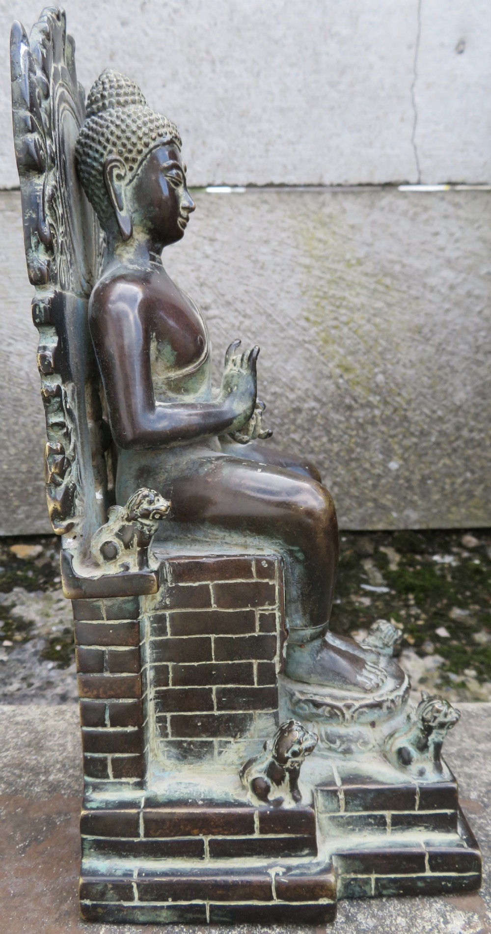 A dark copper alloy sculpture depicting Buddha seated on an ornate throne with legs lowered in the European posture. View XII