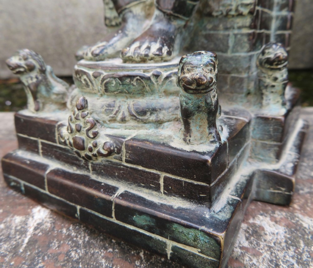 A dark copper alloy sculpture depicting Buddha seated on an ornate throne with legs lowered in the European posture. View XXIII