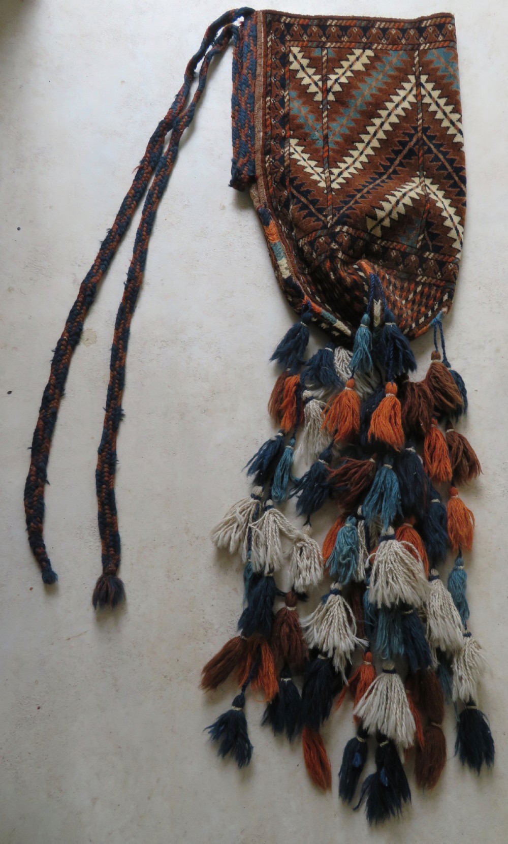 A hand-woven Yomut Turkmen Ok Bash tent-pole bag featuring geometric chevron patterns in rust brown, indigo, and cream wool, finished with braided ties and multi-coloured tassels.