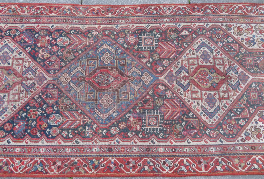 An antique hand-knotted Qashqai wool runner rug featuring repeating geometric diamond medallions in red, blue, and cream tones. View V