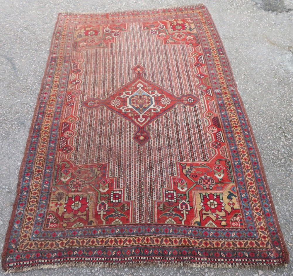 An antique hand-knotted South Persian Khamseh rug featuring a central diamond medallion surrounded by a striped field and geometric borders in shades of terracotta red, navy blue, and cream.