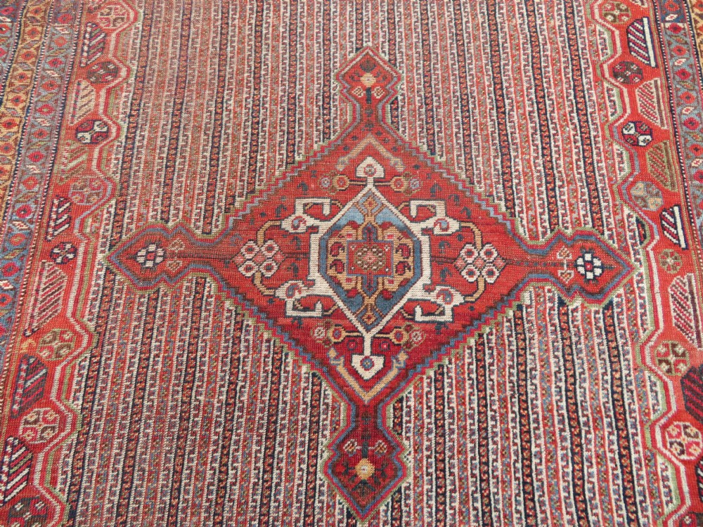 An antique hand-knotted South Persian Khamseh rug featuring a central diamond medallion surrounded by a striped field and geometric borders in shades of terracotta red, navy blue, and cream. View IV