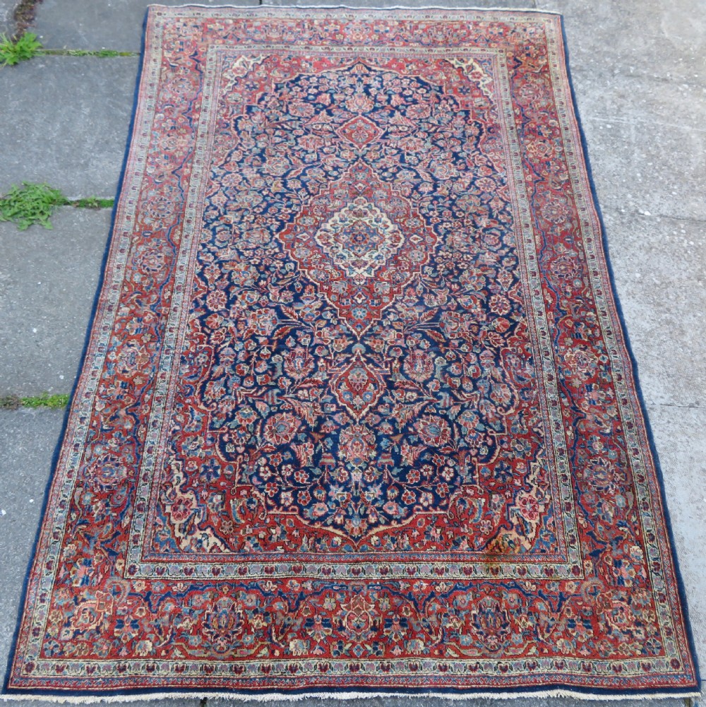 An antique hand-knotted Persian Kashan wool rug featuring a central ivory and red medallion surrounded by dense floral motifs on a dark blue field.