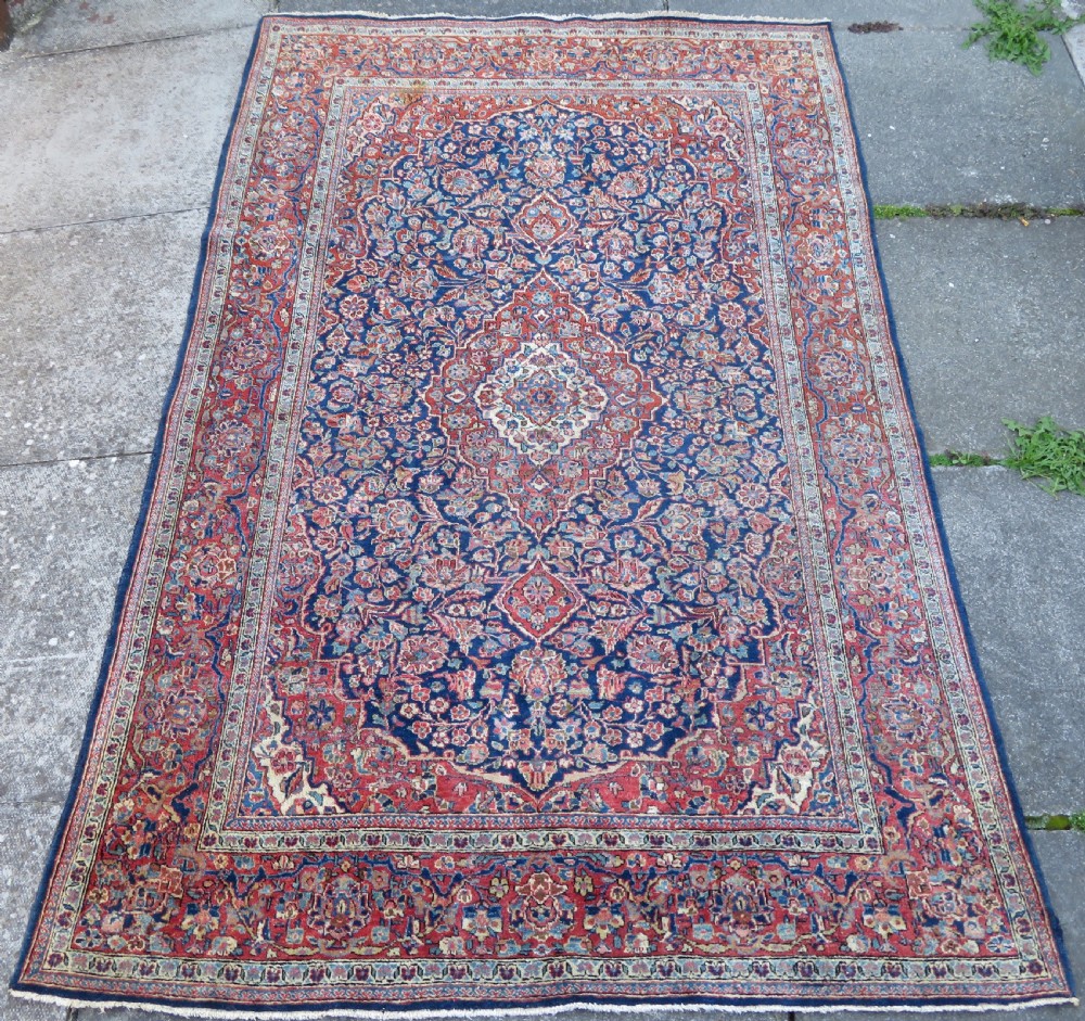 An antique hand-knotted Persian Kashan wool rug featuring a central ivory and red medallion surrounded by dense floral motifs on a dark blue field. View II