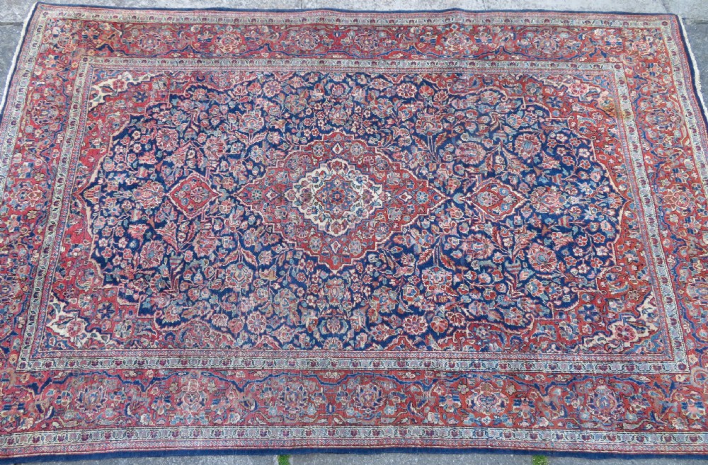 An antique hand-knotted Persian Kashan wool rug featuring a central ivory and red medallion surrounded by dense floral motifs on a dark blue field. View III