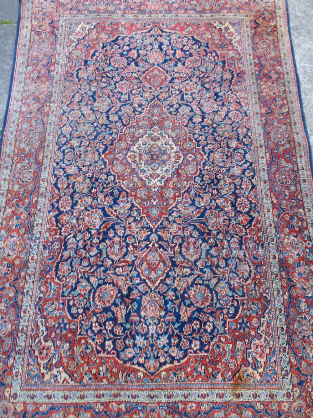 An antique hand-knotted Persian Kashan wool rug featuring a central ivory and red medallion surrounded by dense floral motifs on a dark blue field. View IV
