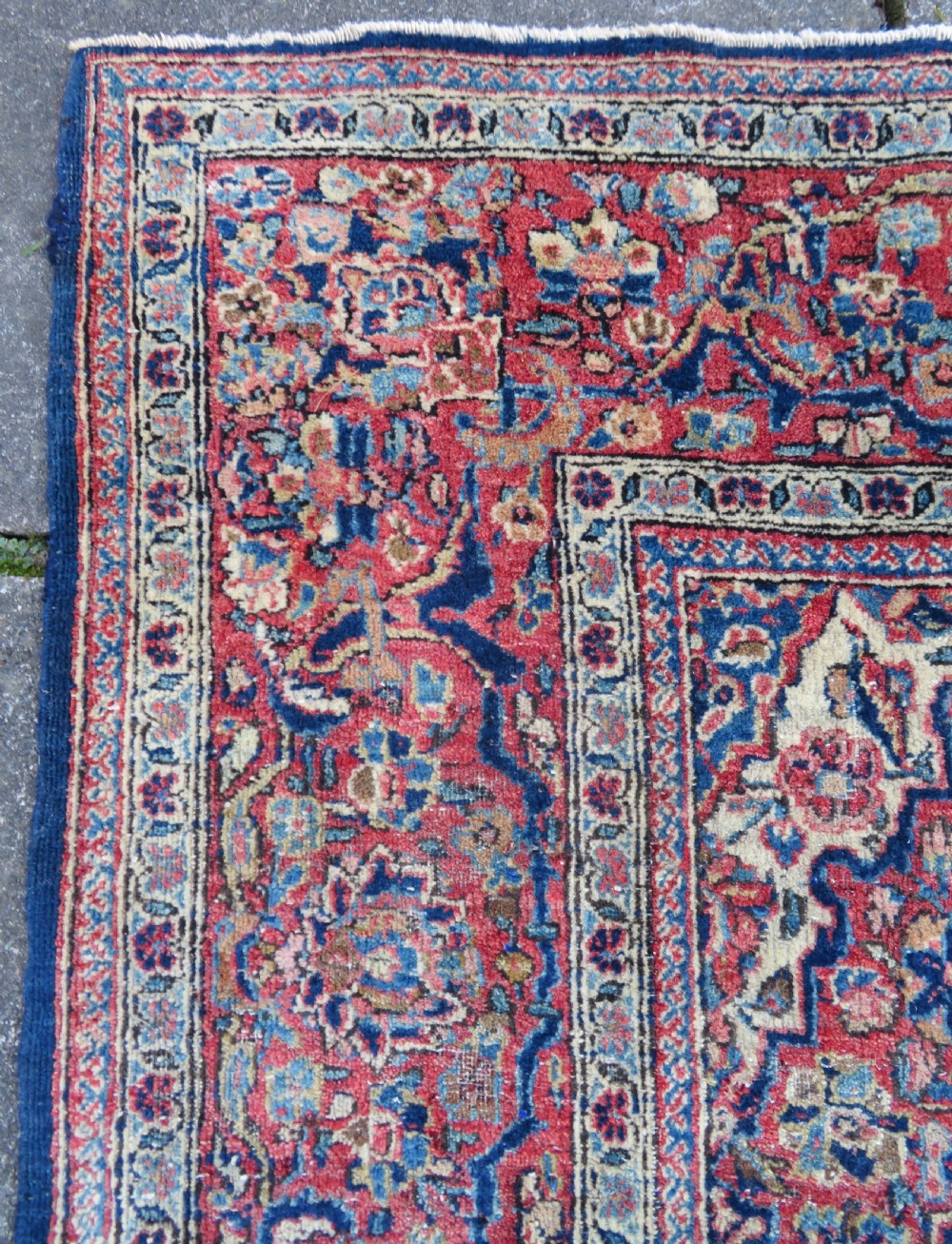 An antique hand-knotted Persian Kashan wool rug featuring a central ivory and red medallion surrounded by dense floral motifs on a dark blue field. View X