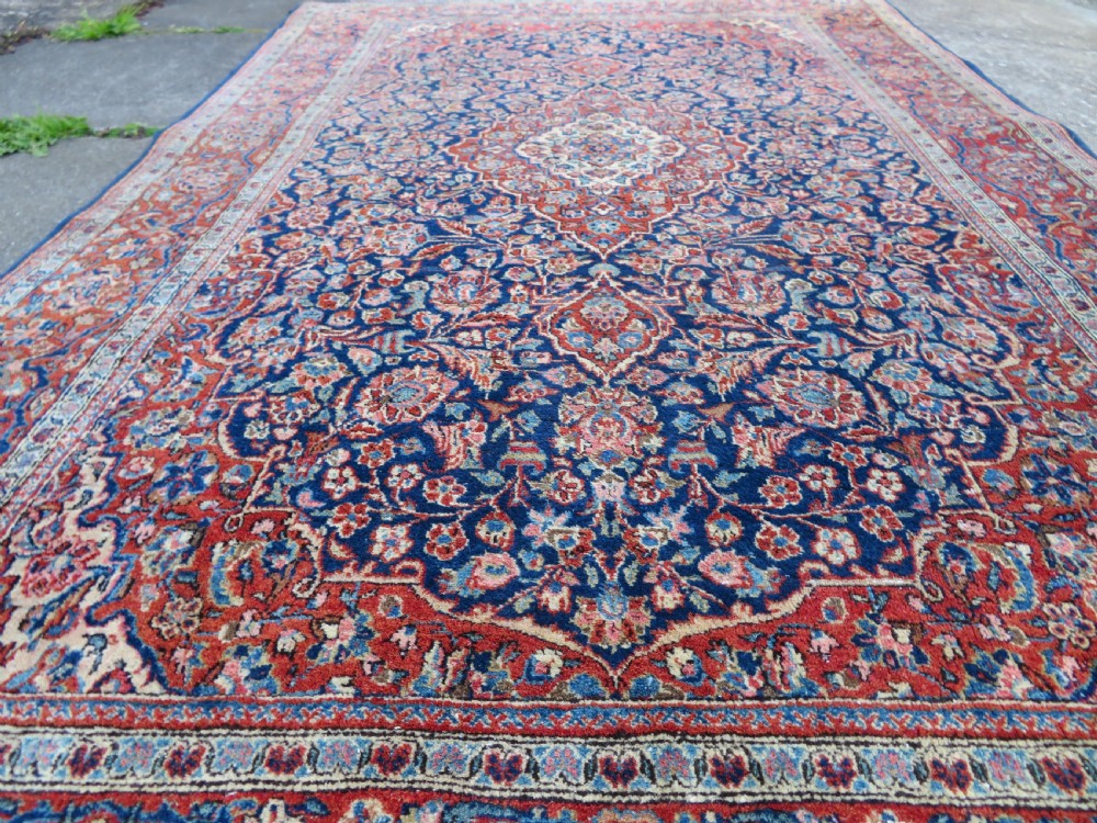 An antique hand-knotted Persian Kashan wool rug featuring a central ivory and red medallion surrounded by dense floral motifs on a dark blue field. View XIV