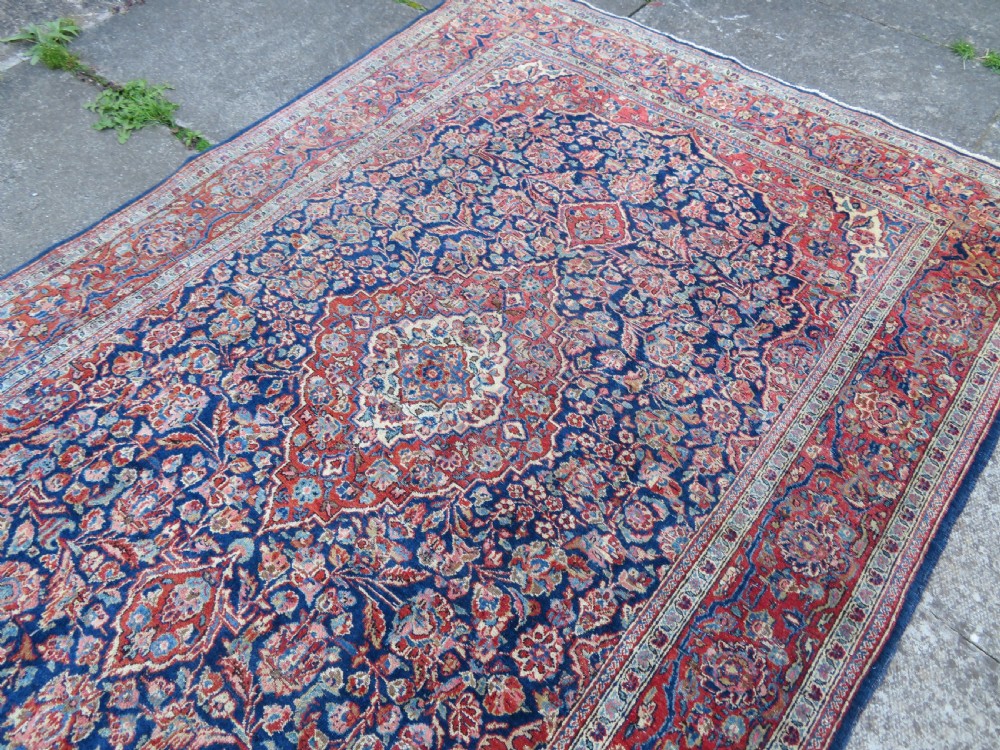 An antique hand-knotted Persian Kashan wool rug featuring a central ivory and red medallion surrounded by dense floral motifs on a dark blue field. View XV