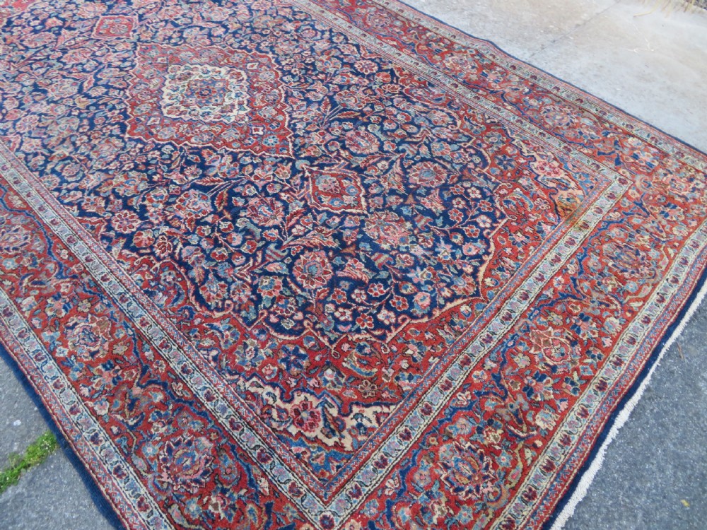 An antique hand-knotted Persian Kashan wool rug featuring a central ivory and red medallion surrounded by dense floral motifs on a dark blue field. View XVI