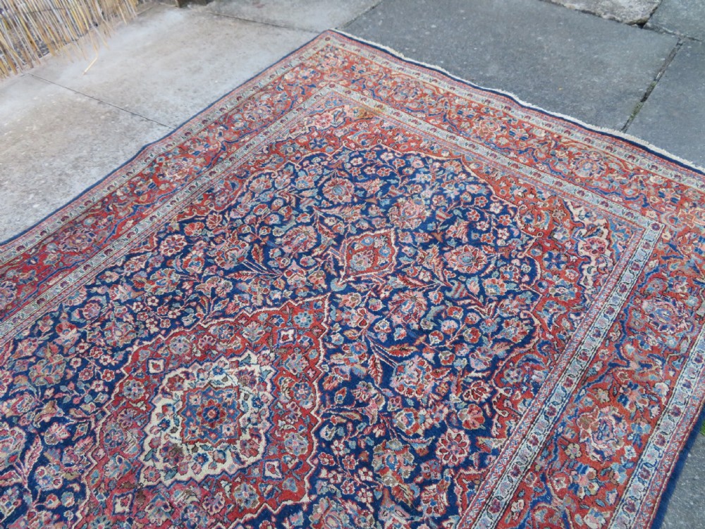 An antique hand-knotted Persian Kashan wool rug featuring a central ivory and red medallion surrounded by dense floral motifs on a dark blue field. View XVII