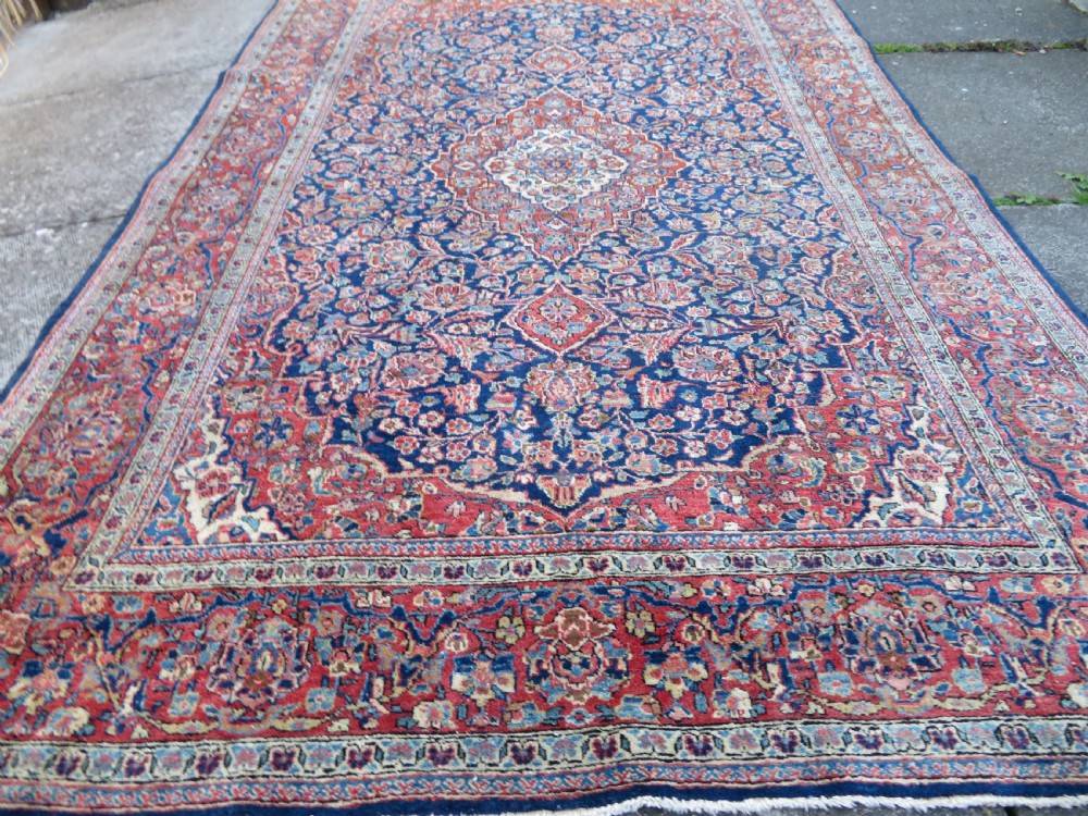 An antique hand-knotted Persian Kashan wool rug featuring a central ivory and red medallion surrounded by dense floral motifs on a dark blue field. View XVIII