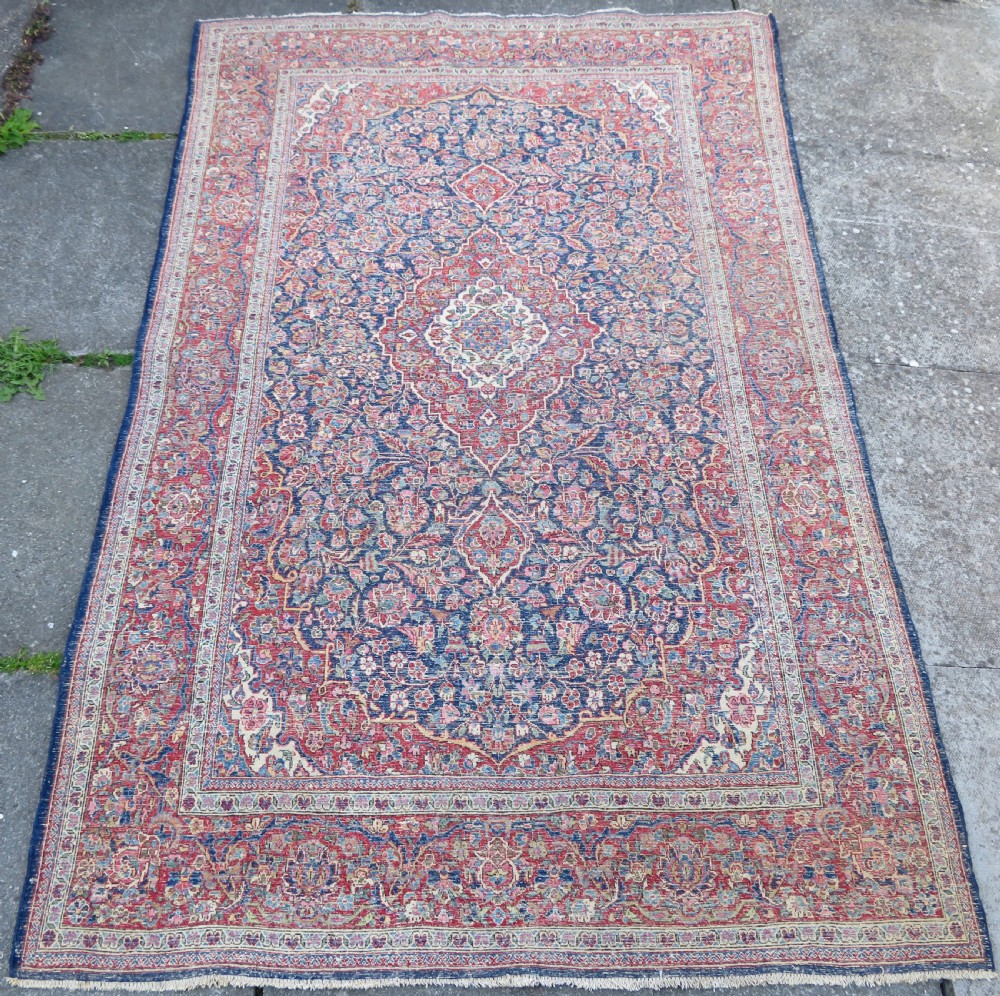 An antique hand-knotted Persian Kashan wool rug featuring a central ivory and red medallion surrounded by dense floral motifs on a dark blue field. View XX