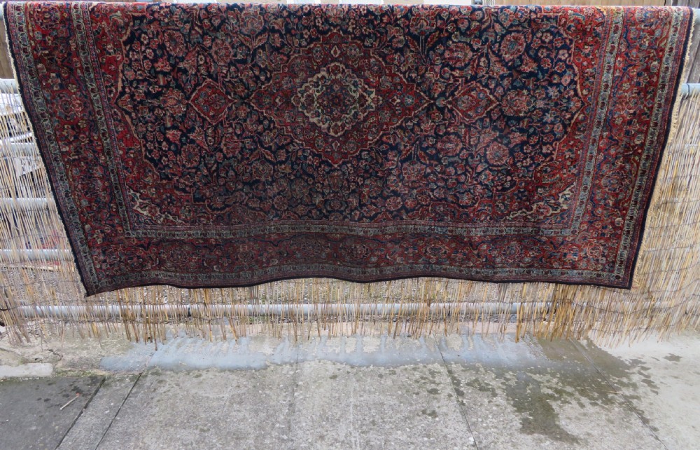 An antique hand-knotted Persian Kashan wool rug featuring a central ivory and red medallion surrounded by dense floral motifs on a dark blue field. View XXIX