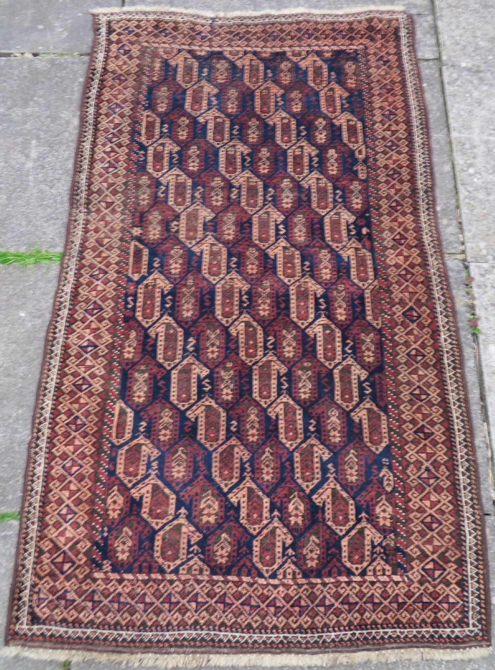 A hand-knotted antique Baluch rug featuring repeating geometric octagonal motifs in shades of dark blue, terracotta, and brown framed by patterned geometric borders.
