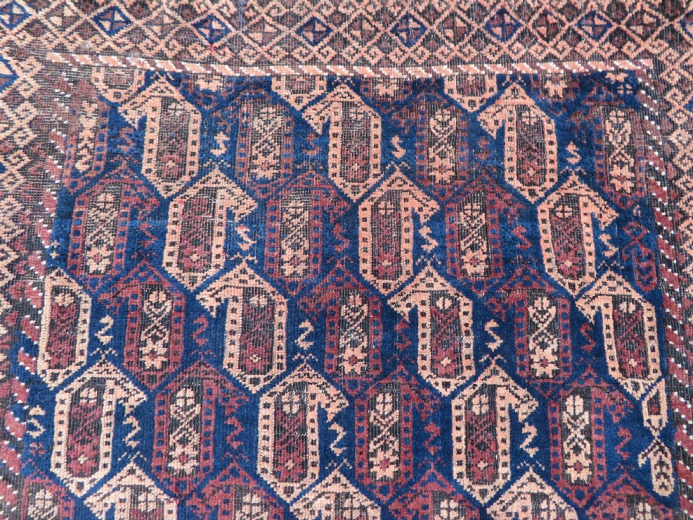 A hand-knotted antique Baluch rug featuring repeating geometric octagonal motifs in shades of dark blue, terracotta, and brown framed by patterned geometric borders. View VI