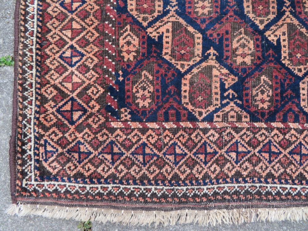 A hand-knotted antique Baluch rug featuring repeating geometric octagonal motifs in shades of dark blue, terracotta, and brown framed by patterned geometric borders. View VII
