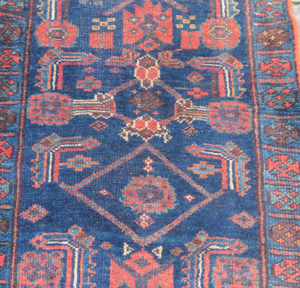 An antique Kurdish wool runner rug featuring geometric and stylized human figures in navy blue, madder red, and ochre with fringed ends. View V