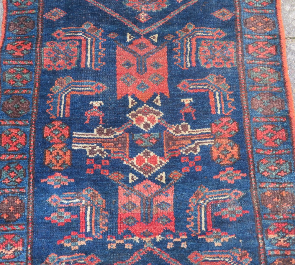 An antique Kurdish wool runner rug featuring geometric and stylized human figures in navy blue, madder red, and ochre with fringed ends. View VI