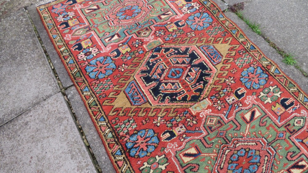 A long, narrow hand-knotted Persian Karaja wool rug runner featuring repeating geometric medallions in navy, blue, and cream against a rust red field. View IX