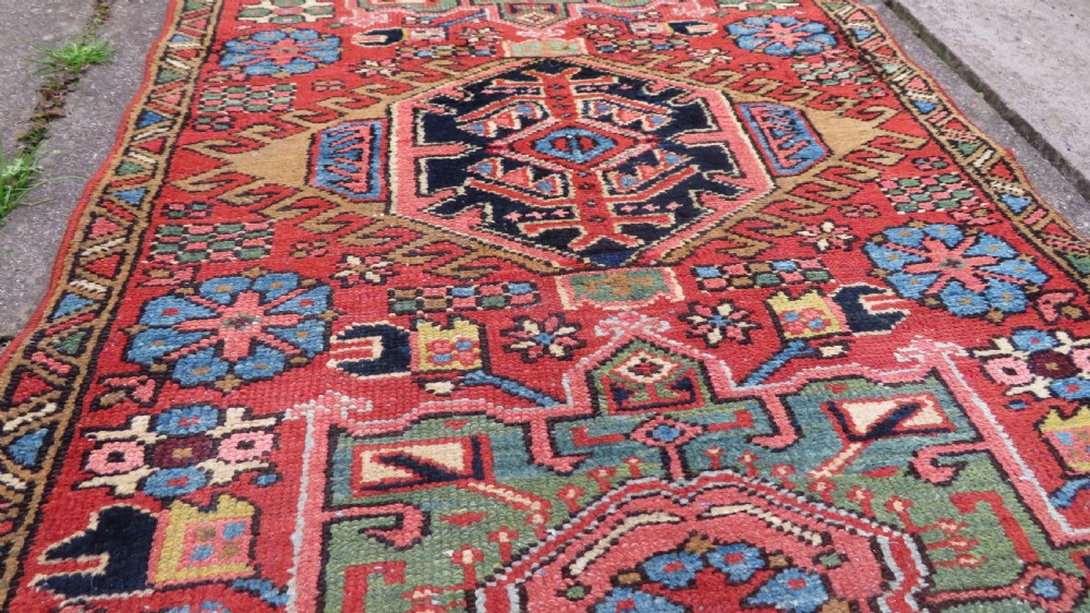 A long, narrow hand-knotted Persian Karaja wool rug runner featuring repeating geometric medallions in navy, blue, and cream against a rust red field. View XV
