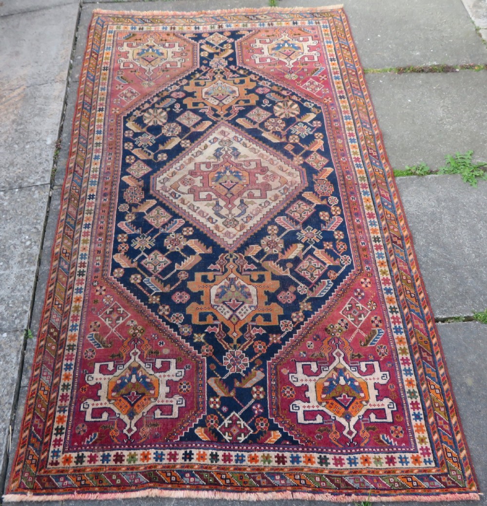 An antique Qashqai hand-knotted wool rug featuring a central diamond medallion surrounded by geometric tribal motifs in shades of navy blue, terracotta red, and cream.