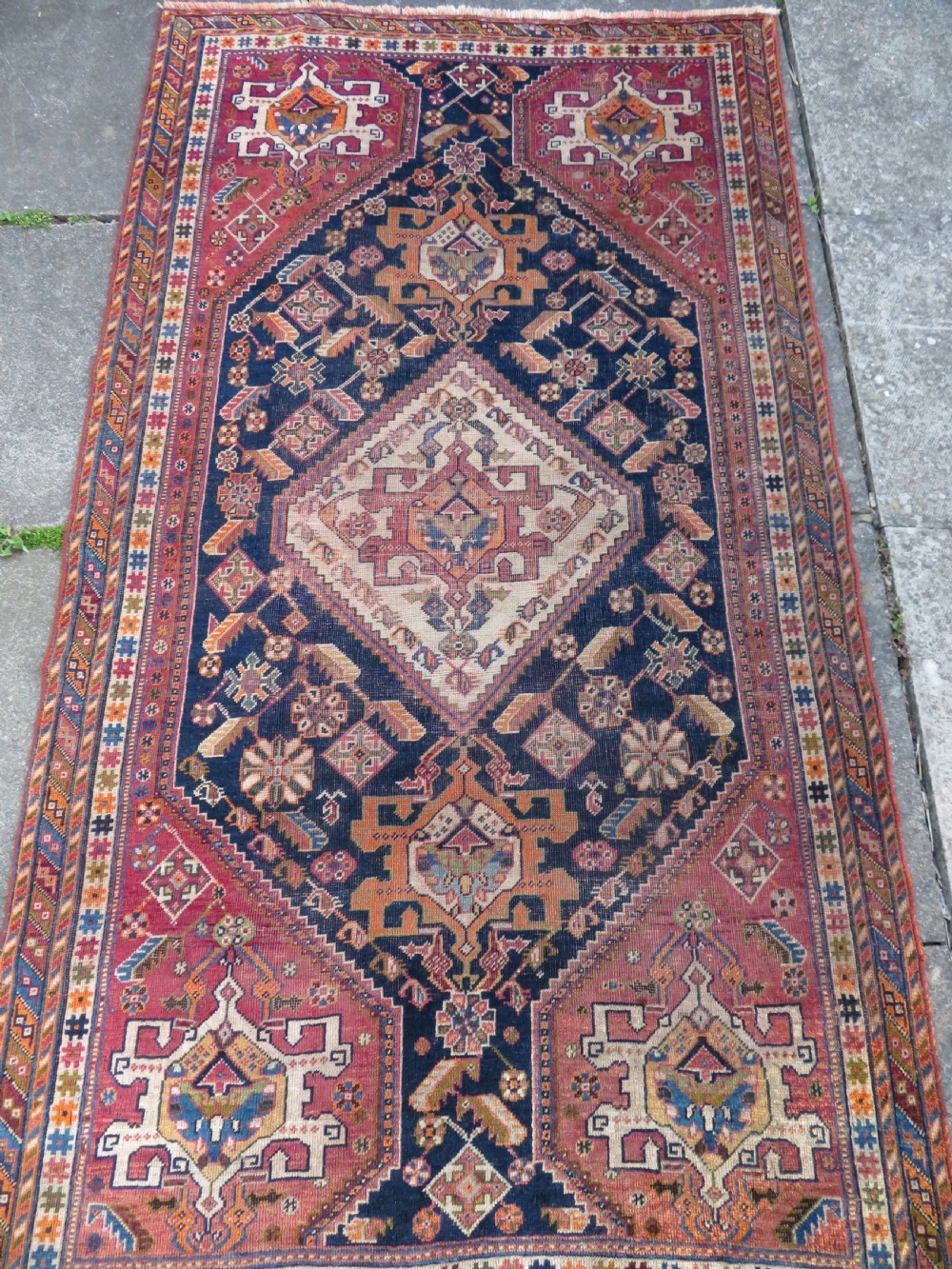 An antique Qashqai hand-knotted wool rug featuring a central diamond medallion surrounded by geometric tribal motifs in shades of navy blue, terracotta red, and cream. View IV