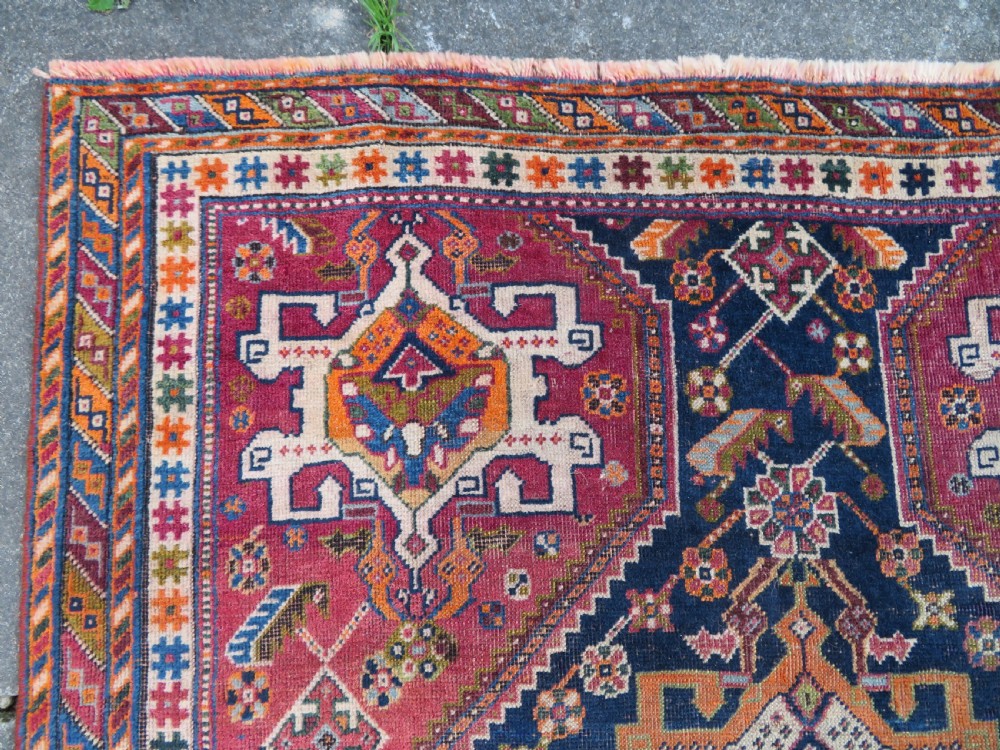 An antique Qashqai hand-knotted wool rug featuring a central diamond medallion surrounded by geometric tribal motifs in shades of navy blue, terracotta red, and cream. View V
