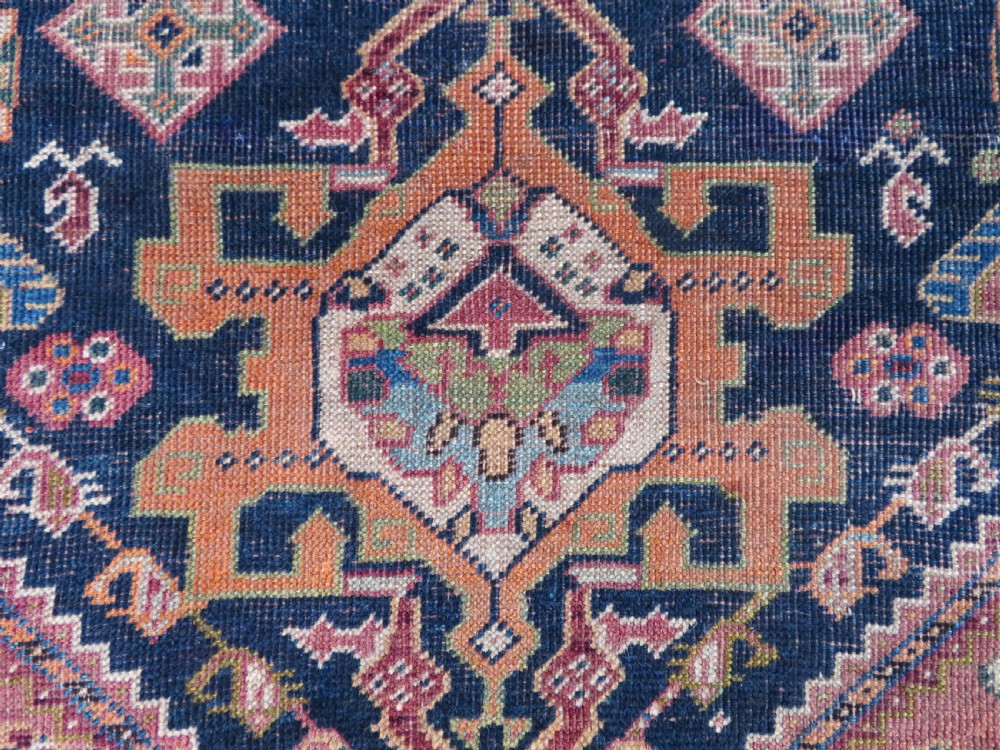 An antique Qashqai hand-knotted wool rug featuring a central diamond medallion surrounded by geometric tribal motifs in shades of navy blue, terracotta red, and cream. View X