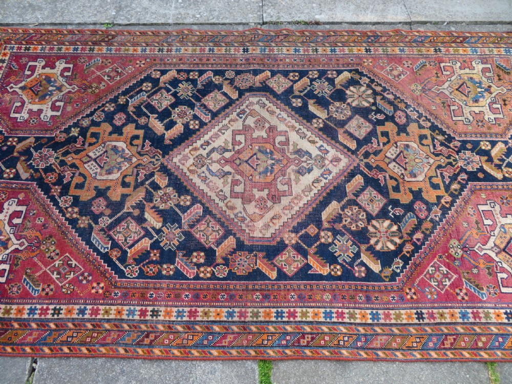 An antique Qashqai hand-knotted wool rug featuring a central diamond medallion surrounded by geometric tribal motifs in shades of navy blue, terracotta red, and cream. View XIX