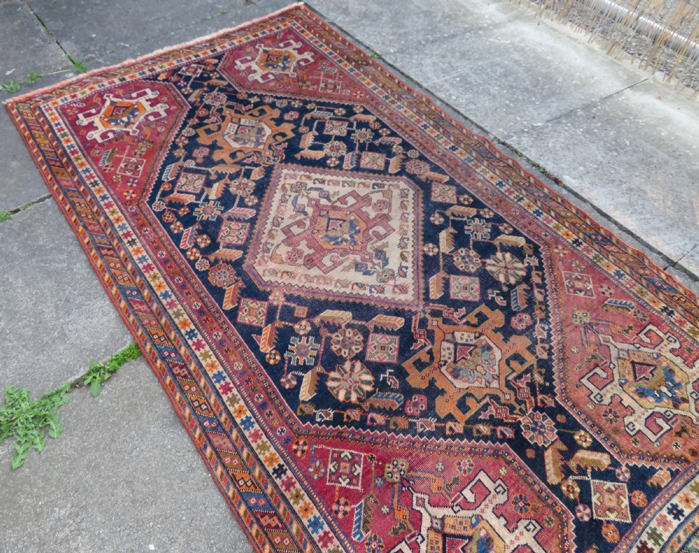 An antique Qashqai hand-knotted wool rug featuring a central diamond medallion surrounded by geometric tribal motifs in shades of navy blue, terracotta red, and cream. View XX