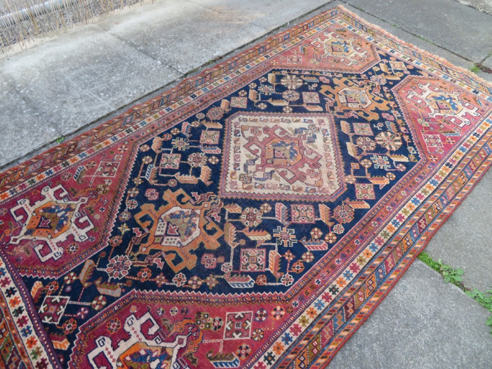 An antique Qashqai hand-knotted wool rug featuring a central diamond medallion surrounded by geometric tribal motifs in shades of navy blue, terracotta red, and cream. View XXI
