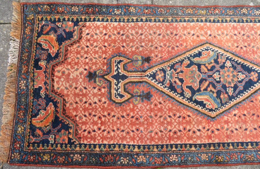 A long, narrow hand-knotted Kurdish runner rug featuring diamond medallions in navy and terracotta on a patterned rust-red field with decorative borders. View III