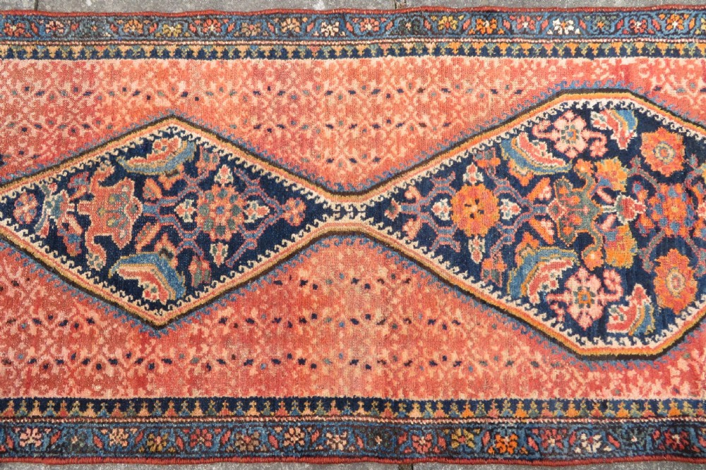 A long, narrow hand-knotted Kurdish runner rug featuring diamond medallions in navy and terracotta on a patterned rust-red field with decorative borders. View IV