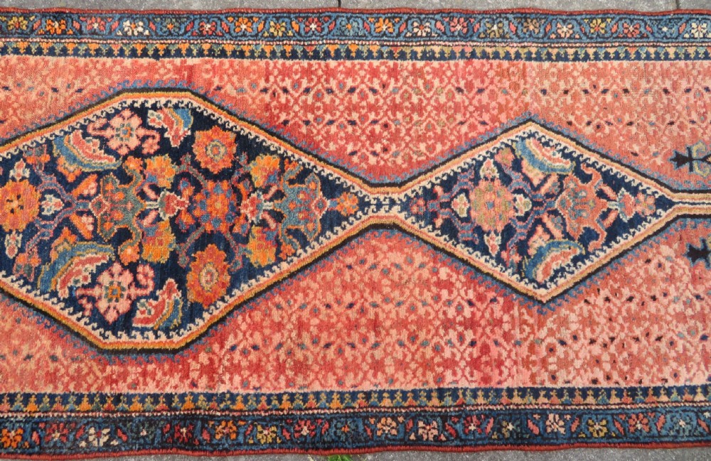 A long, narrow hand-knotted Kurdish runner rug featuring diamond medallions in navy and terracotta on a patterned rust-red field with decorative borders. View V